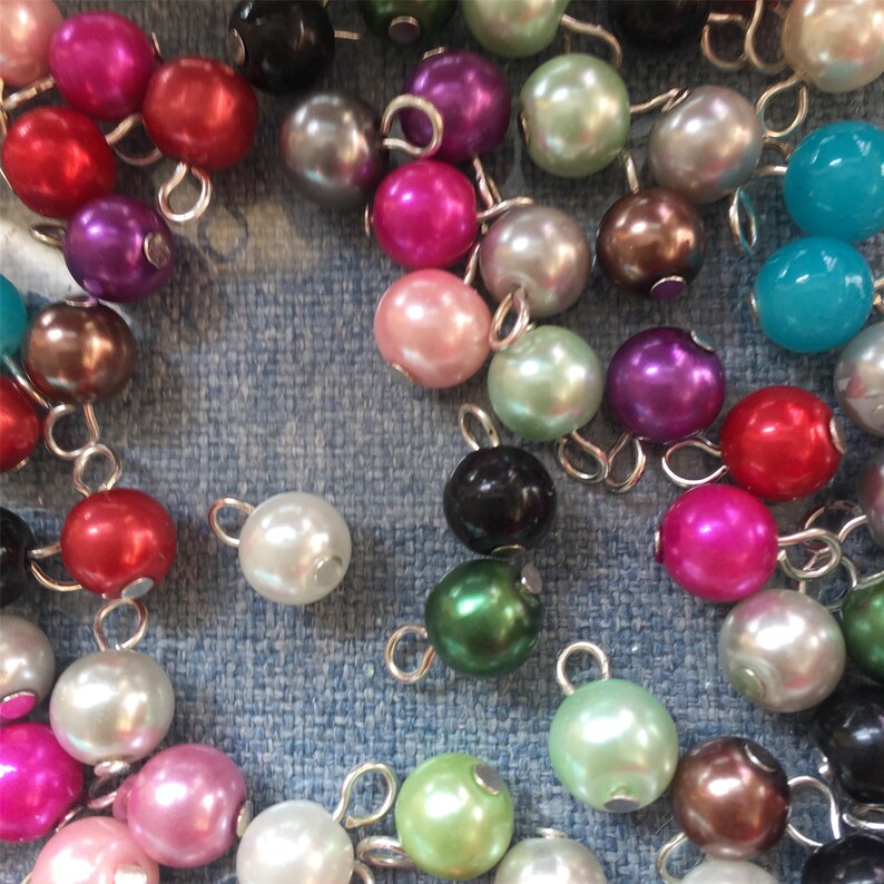 May include: A collection of colourful pearl beads with silver loops. The beads are arranged on a blue fabric surface.