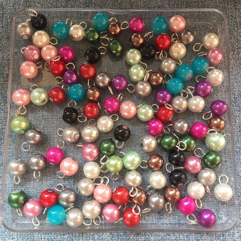 May include: A collection of colourful pearl beads with silver loops. The beads are in various shades of pink, blue, green, and purple. The beads are arranged in a clear plastic container.