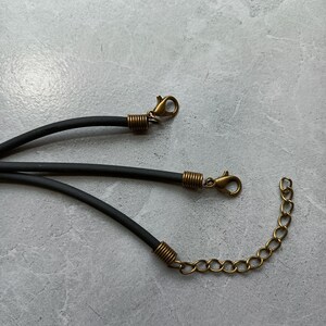 Puede incluir: Tres cordones negros de goma para collares, cada uno con terminales de metal en tono lat&oacute;n antiguo. Dos de los cordones terminan en cierres de mosquet&oacute;n, mientras que el tercero incluye una cadena extensora de metal para ajustar el largo.
