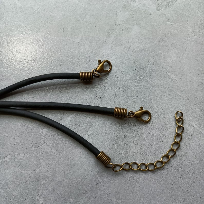 Puede incluir: Tres cordones negros de goma para collares, cada uno con terminales de metal en tono lat&oacute;n antiguo. Dos de los cordones terminan en cierres de mosquet&oacute;n, mientras que el tercero incluye una cadena extensora de metal para ajustar el largo.
