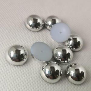 May include: A selection of shiny, silver-coloured, half-sphere beads. Some beads have a flat, light-coloured backing with a small hole. The beads are reflective, showing the surrounding environment. These beads are likely used for crafting or jewellery making.