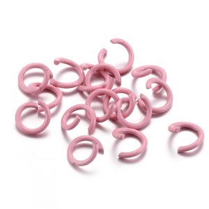 May include: A group of pink metal jump rings. The rings are small and have a slightly open shape. They are ideal for jewelry making or other craft projects.