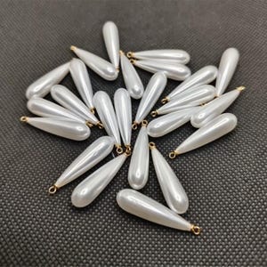 Puede incluir: Una colección de cuentas blancas nacaradas en forma de lágrima con bucles dorados. Estas cuentas están dispersas sobre una superficie texturizada de color gris oscuro. Las cuentas son lisas y brillantes, reflejando la luz.