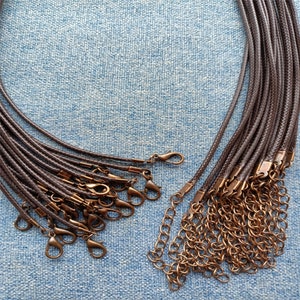 Può includere: Una collezione di collane in cordino intrecciato marrone scuro, ciascuna dotata di una chiusura a moschettone color rame e di una catenella di estensione in metallo abbinato all'estremità per regolare la lunghezza, disposte in un fascio su una superficie blu testurizzata.