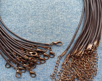 Puede incluir: Una colección de collares de cordón trenzado de color marrón oscuro, cada uno con un cierre de mosquetón de metal en tono cobre y una cadena extensora ajustable. Los cordones están dispuestos en un manojo, mostrando su textura uniforme.