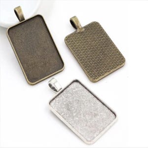 May include: Three silver and bronze rectangular pendant trays with a loop at the top for hanging. The trays are blank and ready for crafting.
