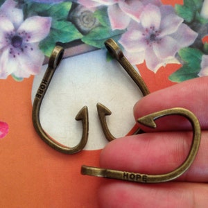 May include: Close-up of several bronze-coloured metal hook clasps. The clasps are curved with an arrow-shaped end and the word "HOPE" stamped on the side. The clasps are on a floral background.