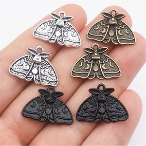 Puede incluir: Cinco abalorios de polilla plateados, negros y bronce con un diseño de luna creciente y estrella. Los abalorios están en una mano.