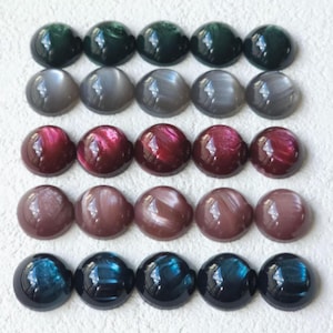 May include: A collection of twenty cabochon gemstones in a variety of colors. The gemstones are arranged in five rows of four. Colors include dark green, gray, red, brown, and blue. Each stone has a smooth, rounded surface.