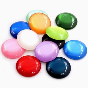 Può includere: Una collezione di cabochon rotondi e lucidi in vari colori, tra cui rosso, rosa, bianco, viola, blu, verde e arancione. Queste pietre lisce e lucide hanno un effetto occhio di gatto, creando un aspetto scintillante. I cabochon sono disposti su una superficie bianca.