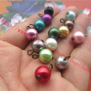 May include: Assortment of pearl beads with metal loops in various colours, including pink, green, white, and grey. The beads are approximately 1 cm in diameter.