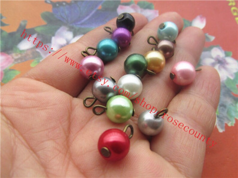 May include: Assortment of pearl beads with metal loops in various colours, including pink, green, white, and grey. The beads are approximately 1 cm in diameter.