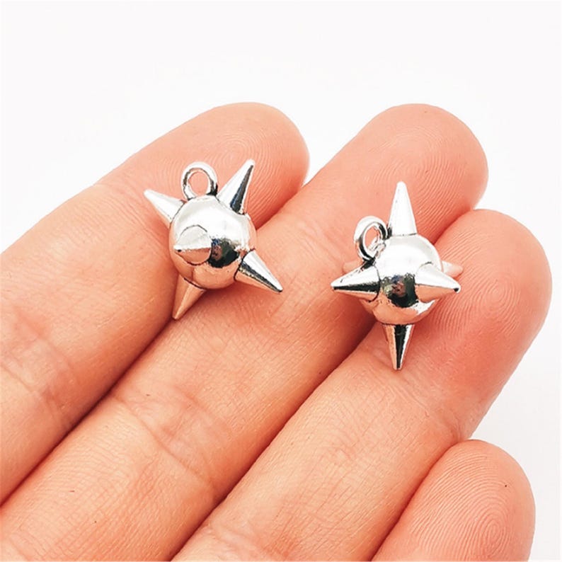 May include: Two silver-colored, spiky charms with a loop at the top for hanging. Each charm has a central sphere with multiple conical spikes radiating outwards. The charms are held in a person's hand against a white background.