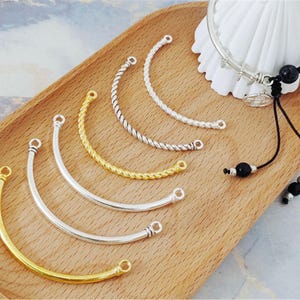 Puede incluir: Una colección de componentes de joyería curvos en tonos plateados y dorados, con diseños lisos y trenzados. Las piezas están dispuestas sobre una bandeja de madera, con una pulsera y elementos decorativos en el fondo.