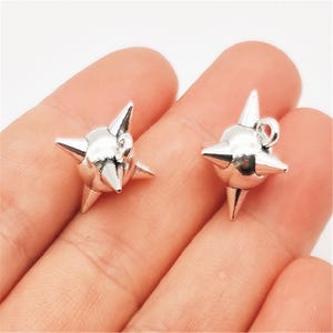 May include: Two silver-colored starburst charms with pointed spikes. Each charm has a small loop for attaching to jewelry. The charms are held in a person's hand.