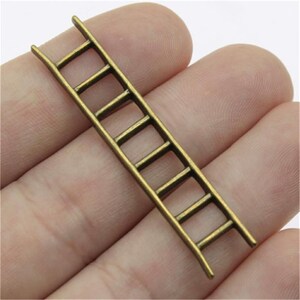 Può includere: Un piccolo ciondolo a forma di scala color bronzo, con sette pioli. La scala è in metallo e ha un design leggermente curvo. Il ciondolo è tenuto in mano, mostrando le sue dimensioni e i dettagli. Ideale per la creazione di gioielli o lavori artigianali.
