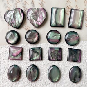 May include: An assortment of iridescent cabochons in heart, rectangular, square, circular, and oval shapes. The cabochons display a swirling pattern of green, purple, and black, with a glossy finish. These decorative pieces are suitable for jewellery making or crafts.