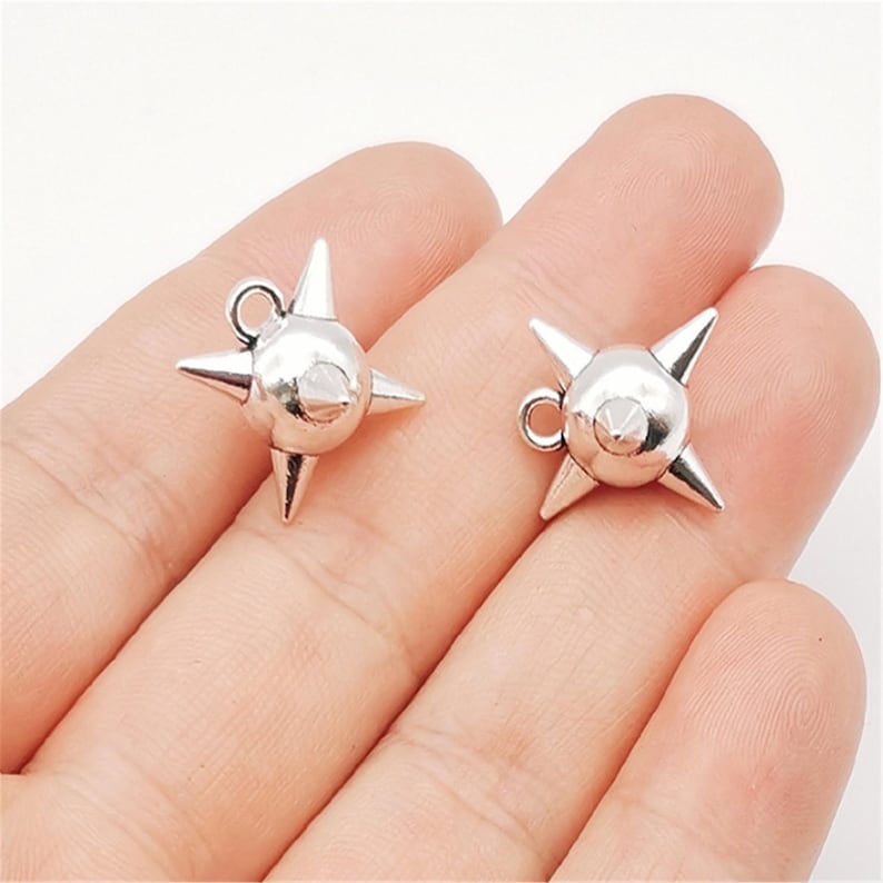 May include: Two silver-colored starburst charms with a central sphere and four pointed spikes. Each charm has a small loop for attaching to jewelry or other crafts. The charms are shown against a light background.