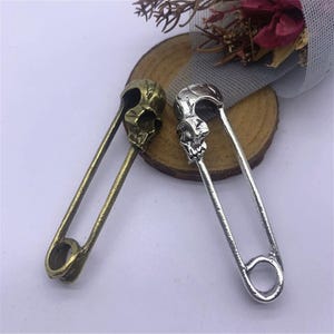 Può includere: Due spille di sicurezza con teste a forma di teschio. Una spilla è argentata e l'altra è di bronzo.