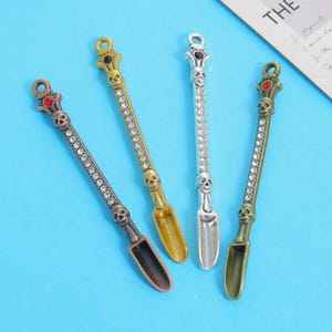 Pode incluir: Quatro colheres decorativas com desenhos de caveiras e cristais. As colheres têm diferentes acabamentos metálicos: bronze, ouro e prata. Cada colher apresenta uma caveira no topo e ao longo do cabo, com uma pequena concha na extremidade.