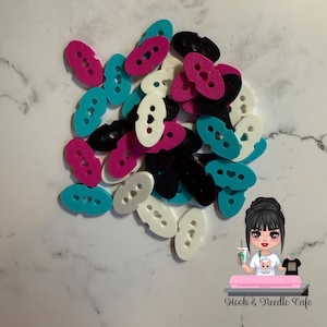 Puede incluir: Un conjunto de botones ovalados en varios colores, incluyendo rosa, turquesa, negro y blanco. Cada botón tiene dos agujeros y un recorte en forma de corazón. Los botones están dispersos sobre una superficie blanca.
