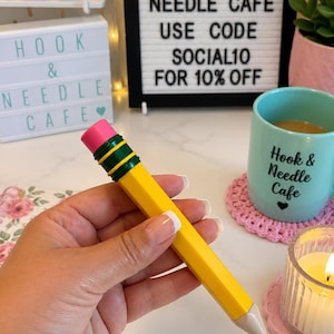 May include: A yellow pencil-shaped pen with a pink eraser, a dark green metal band, and a light tan tip. The pen has a hexagonal body and a fine black ink point. The design mimics a classic wooden pencil with a bright yellow barrel and a pink top.