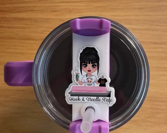 May include: Purple travel mug with a clear lid and a straw. The mug has a sticker with a cartoon illustration of a woman holding a cup of coffee and a sewing machine. The sticker also says "Hook & Needle Cafe".