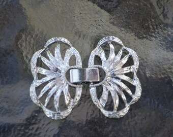May include: A silver metal clasp with a decorative design resembling a flower. The clasp is made of two pieces that connect in the middle.