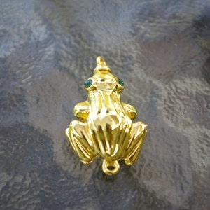 May include: A gold-colored frog charm with green gem eyes. The frog is sitting on a gray surface.