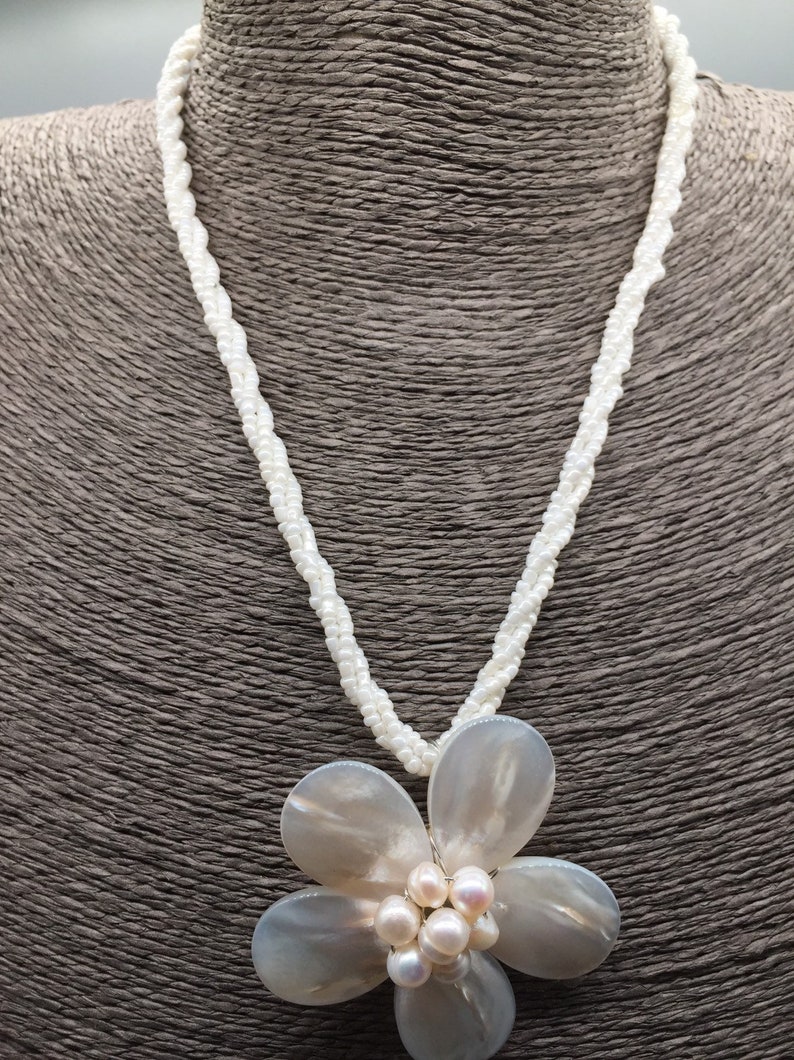 Pode incluir: Um colar de p&eacute;rolas brancas com um grande pingente de flor de concha branca. A flor tem um grupo de p&eacute;rolas no centro.