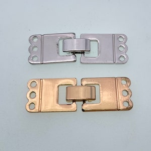 Puede incluir: Dos cierres metálicos, uno plateado y otro dorado, con un diseño decorativo y tres bucles a cada lado. Los cierres se utilizan para conectar piezas de joyería.