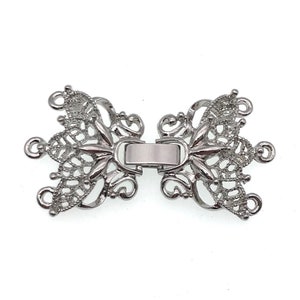 Puede incluir: Cierre de filigrana plateado con un diseño decorativo. El cierre está hecho de dos piezas que se conectan en el medio.