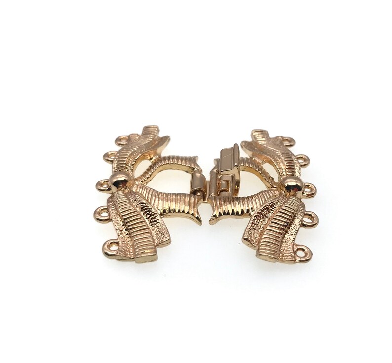 K&ouml;nnte beinhalten: Goldfarbener Metallverschluss mit dekorativem Design und mehreren Schlaufen zum Anbringen von Schmuck oder anderen Gegenst&auml;nden.