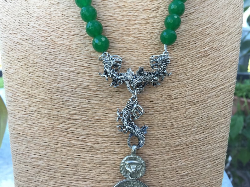 May include: A green jade bead necklace with a silver dragon pendant. The pendant features two dragons facing each other with a third dragon hanging below.