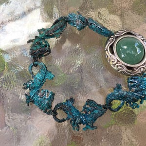May include: A turquoise blue bracelet with a silver charm featuring a green stone. The bracelet is made of metal and features a design of multiple seahorses.