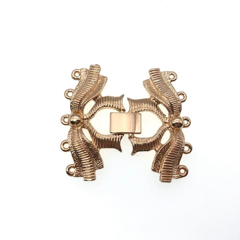 K&ouml;nnte beinhalten: Goldfarbener Metallverschluss mit dekorativem Design. Der Verschluss hat auf jeder Seite vier Schlaufen zum Anbringen von Schmuck.