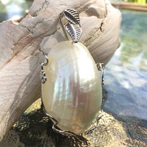 Puede incluir: Un colgante de concha blanca iridiscente con un dise&ntilde;o de hoja de plata. El colgante tiene forma ovalada y tiene una anilla de plata.