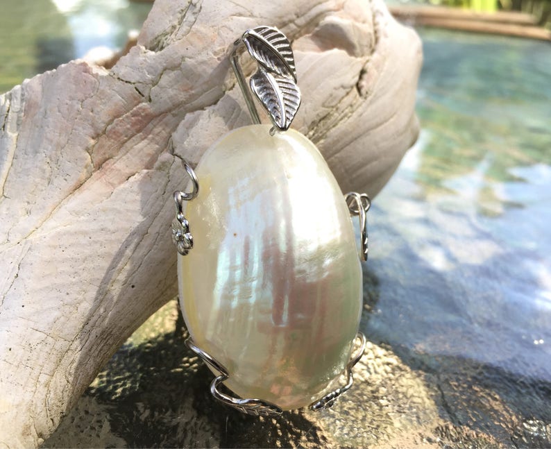 Puede incluir: Un colgante de concha blanca iridiscente con un dise&ntilde;o de hoja de plata. El colgante tiene forma ovalada y tiene una anilla de plata.