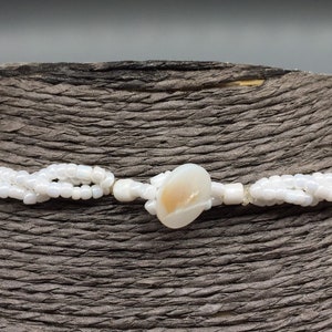 Pode incluir: Um colar de contas brancas com uma &uacute;nica conta grande de concha de cor branco-creme no centro.