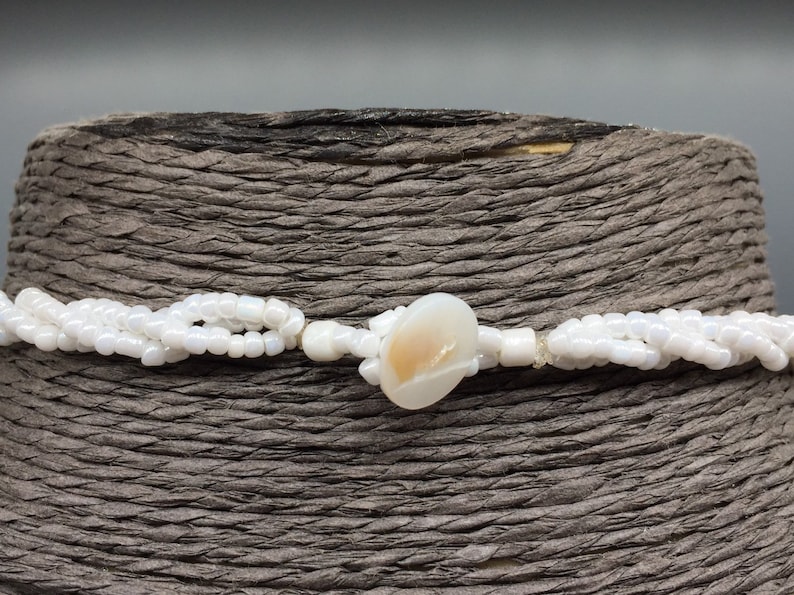 Pode incluir: Um colar de contas brancas com uma &uacute;nica conta grande de concha de cor branco-creme no centro.