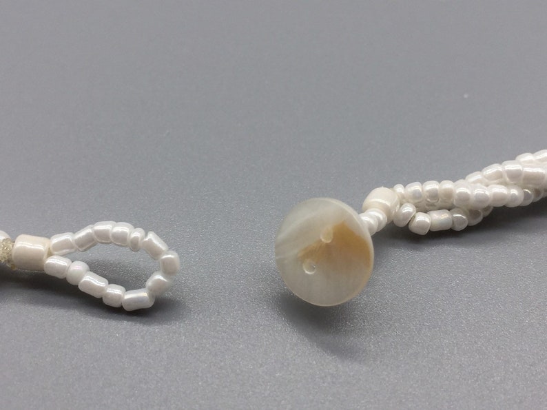 Pode incluir: Um colar de contas brancas com um fecho de bot&atilde;o branco. As contas s&atilde;o pequenas e redondas.