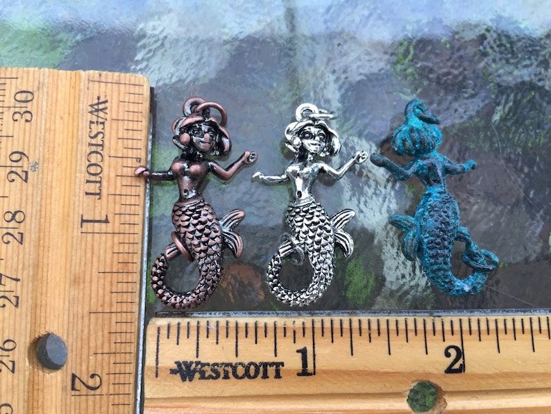 May include: Three mermaid charms in different colors: copper, silver, and blue. The charms are about 1 inch tall and are shown next to a ruler.