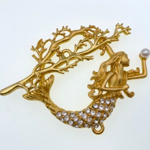 May include: Gold-tone mermaid charm with a pearl, surrounded by a gold-tone branch design. The mermaid's tail is adorned with clear rhinestones.