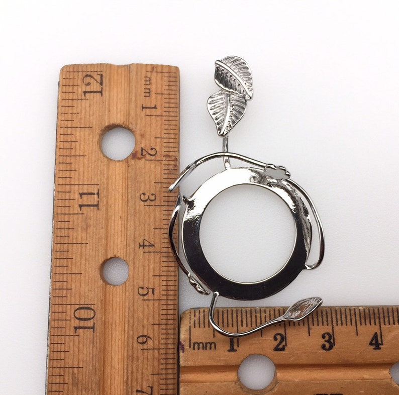 Puede incluir: Pendiente de metal plateado con un marco circular y un dise&ntilde;o de hojas. El pendiente mide aproximadamente 5 cm de di&aacute;metro y se mide con una regla.