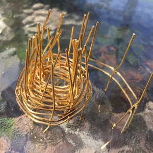 May include: A gold wire sculpture with a circular base and a tall, spiky top. The sculpture is made of thin, interwoven wires that create a delicate and intricate design.