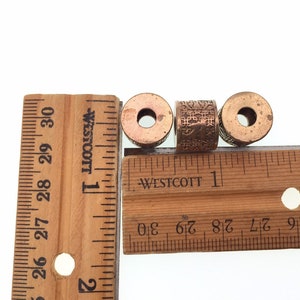 Puede incluir: Tres cuentas de metal de color cobre con dise&ntilde;os intrincados. Las cuentas tienen aproximadamente 2,5 cm de di&aacute;metro y est&aacute;n alineadas en una regla de madera.