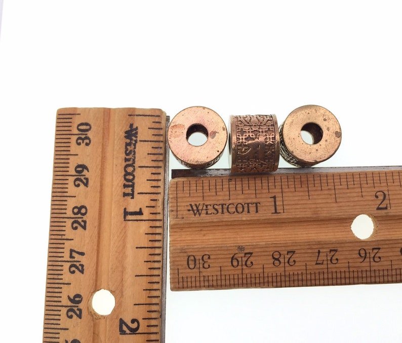 Puede incluir: Tres cuentas de metal de color cobre con dise&ntilde;os intrincados. Las cuentas tienen aproximadamente 2,5 cm de di&aacute;metro y est&aacute;n alineadas en una regla de madera.