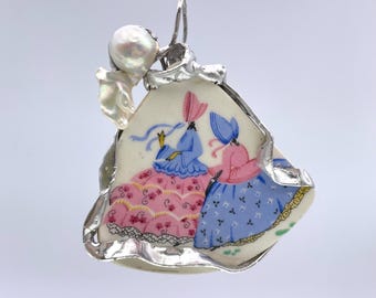 May include: A pendant featuring a porcelain shard with a hand-painted design of two women in dresses and bonnets. The shard is encased in a silver metal frame and adorned with a pearl. The women's dresses are pink and blue.