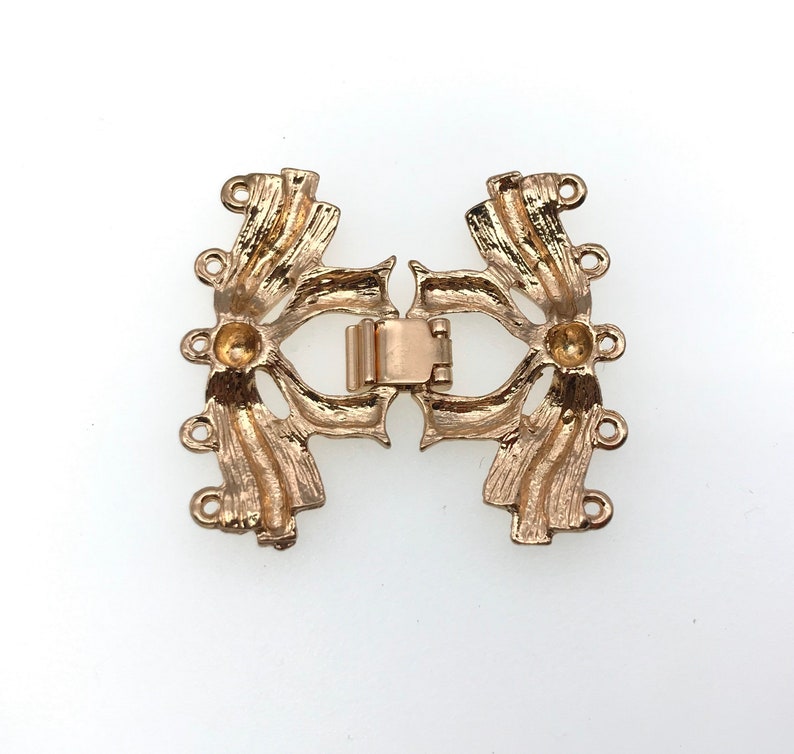 K&ouml;nnte beinhalten: Goldfarbener Metallverschluss mit dekorativem Design und vier Schlaufen auf jeder Seite. Der Verschluss ist offen und bereit, an eine Halskette oder ein Armband befestigt zu werden.