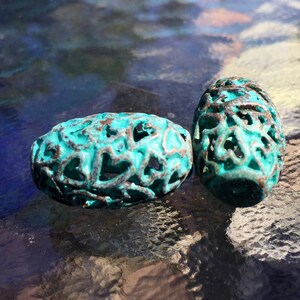 May include: Two teal blue oval-shaped beads with a decorative, intricate, openwork design. The beads are made of metal and have a distressed, aged look.